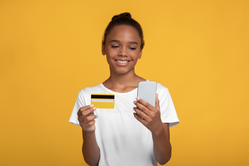 lächelndes Mädchen vor einem gelben Hintergrund mit einer Girokarte und Smartphone in der Hand, auf der Suche nach dem passenden Girokonto für Auszubildende